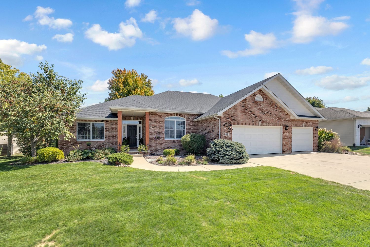 Brick ranch-style home with two-car garage, manicured lawn, and mature trees under blue sky.