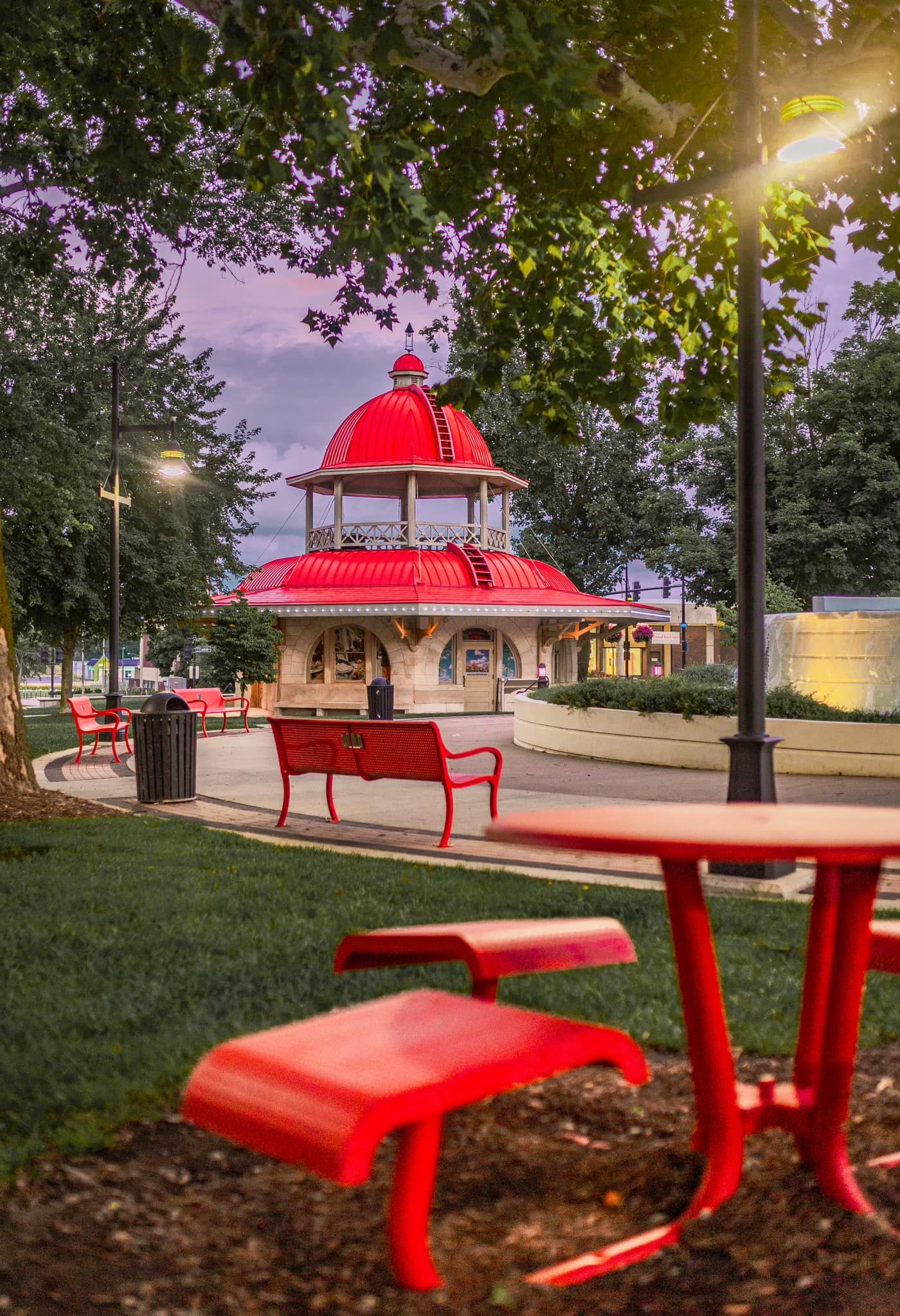 Red picnic table and benches near a charming red-domed gazebo pavilion in a peaceful evening park.