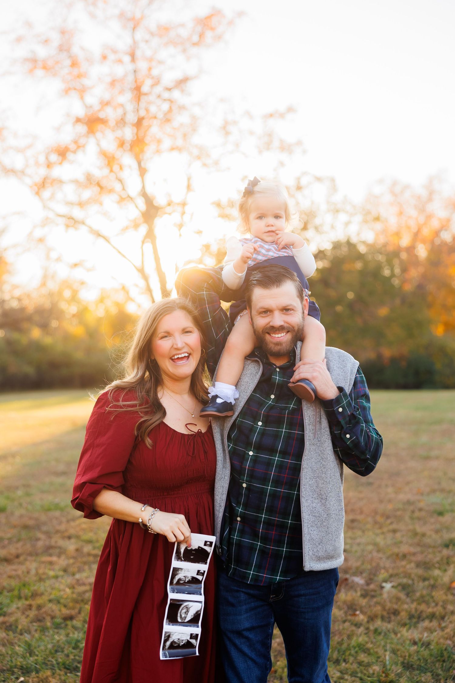 Happy couple with toddler on shoulders holding ultrasound photos in golden hour outdoor setting.