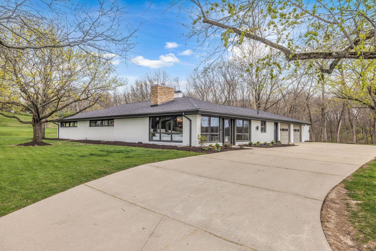 White mid-century modern ranch home with long driveway, attached garage, and wooded backyard setting.