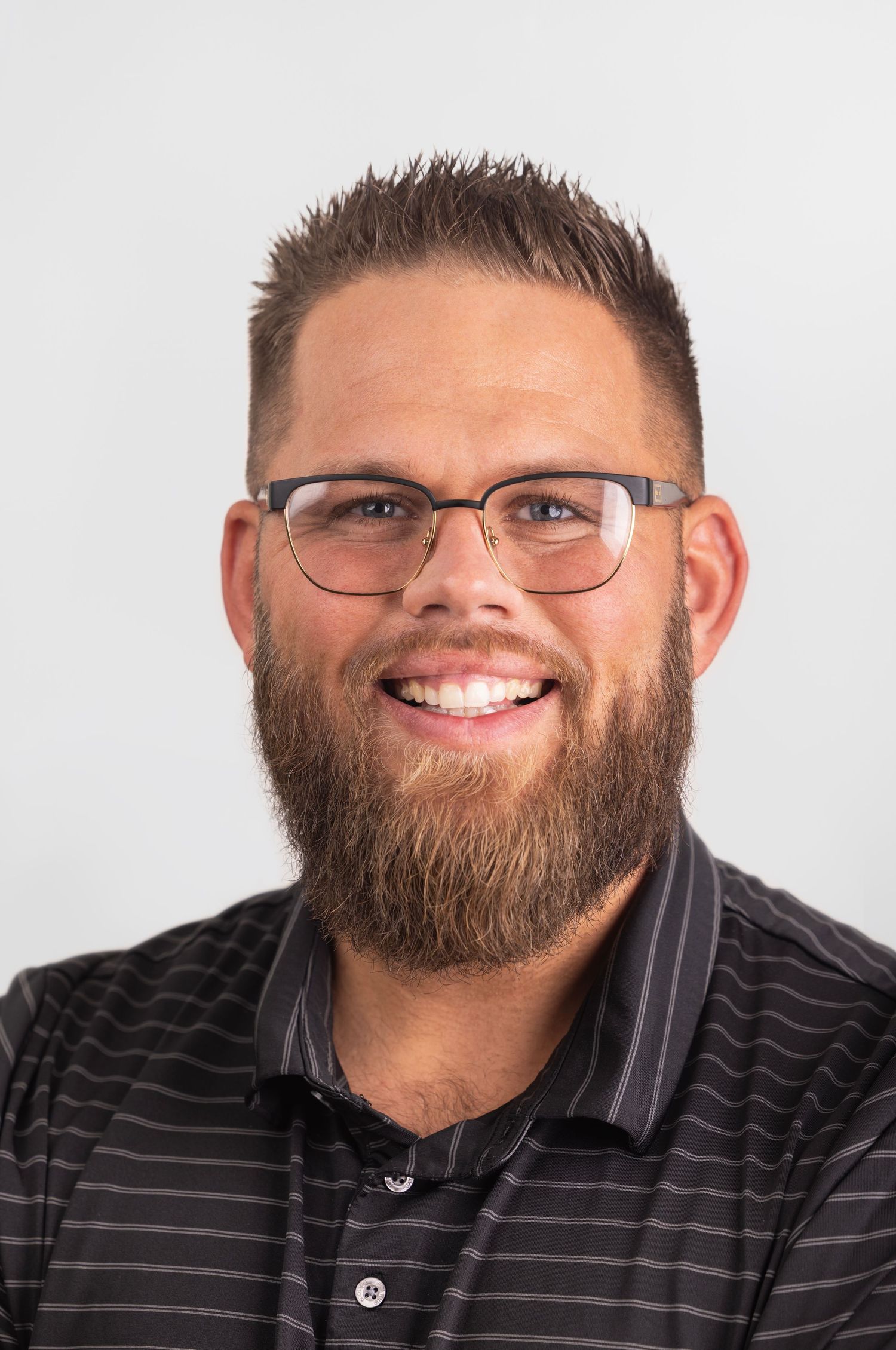 Professional headshot of a smiling bearded man with glasses wearing a striped polo shirt.