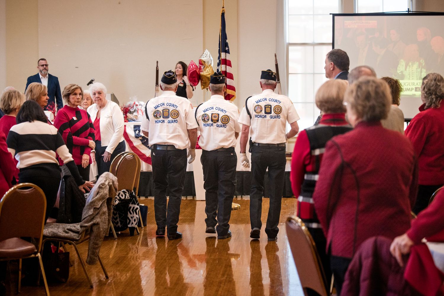 Veterans in matching white shirts carry flags at a formal indoor ceremony with seated attendees in red attire.