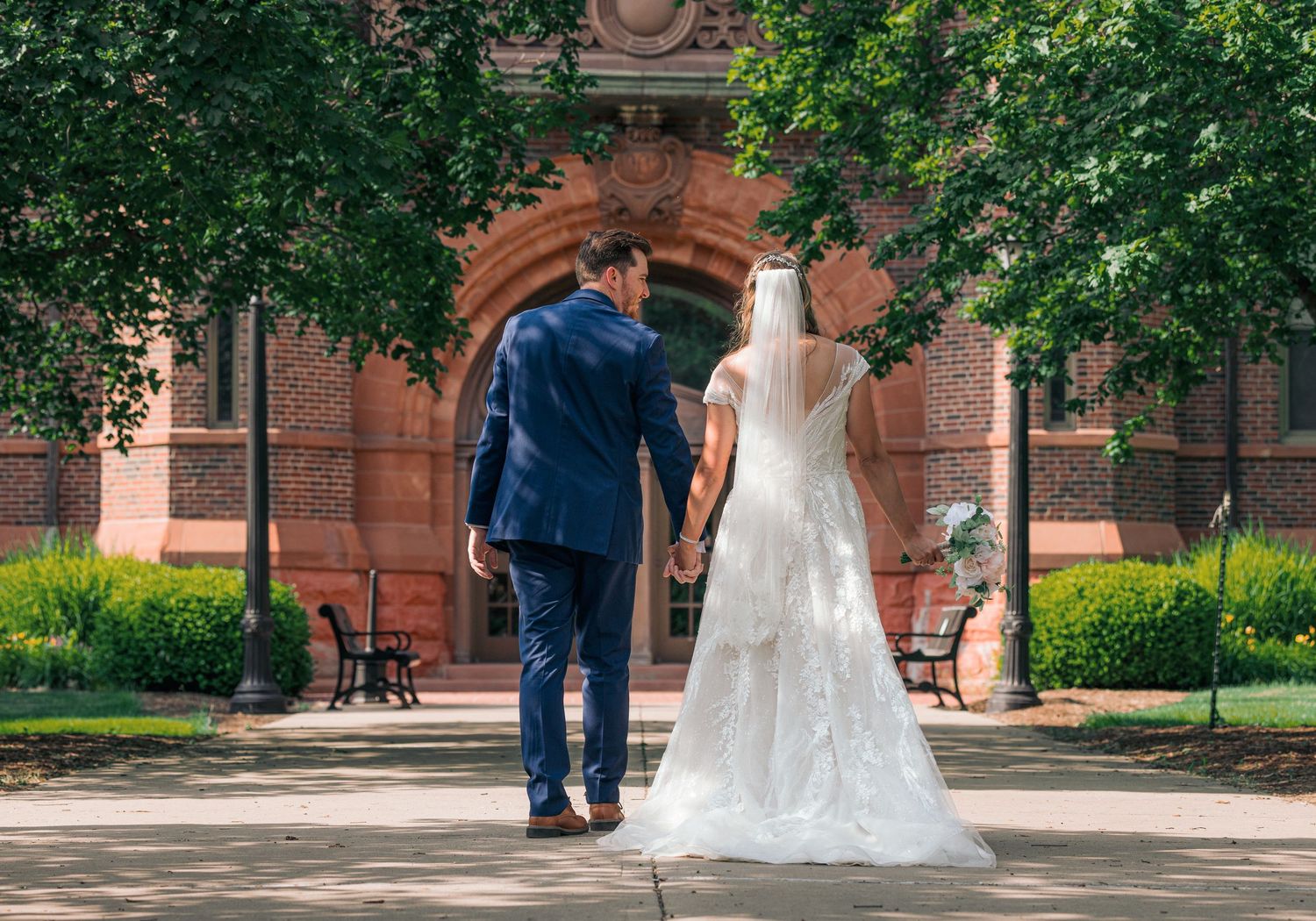 Bride and groom holding hands walking toward a red brick arched building surrounded by lush greenery.