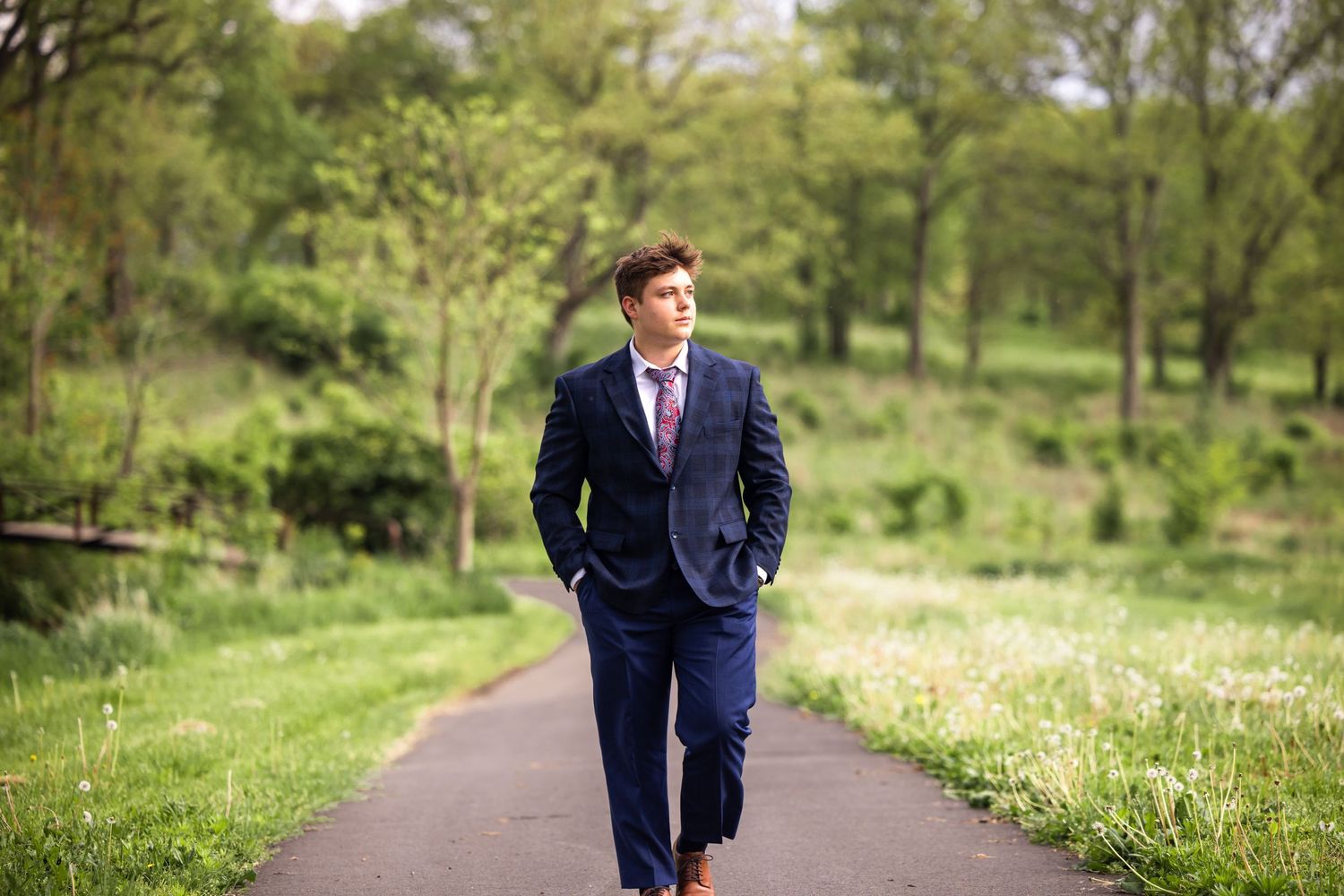 Young man in navy suit walking confidently along a tree-lined path in a green park.