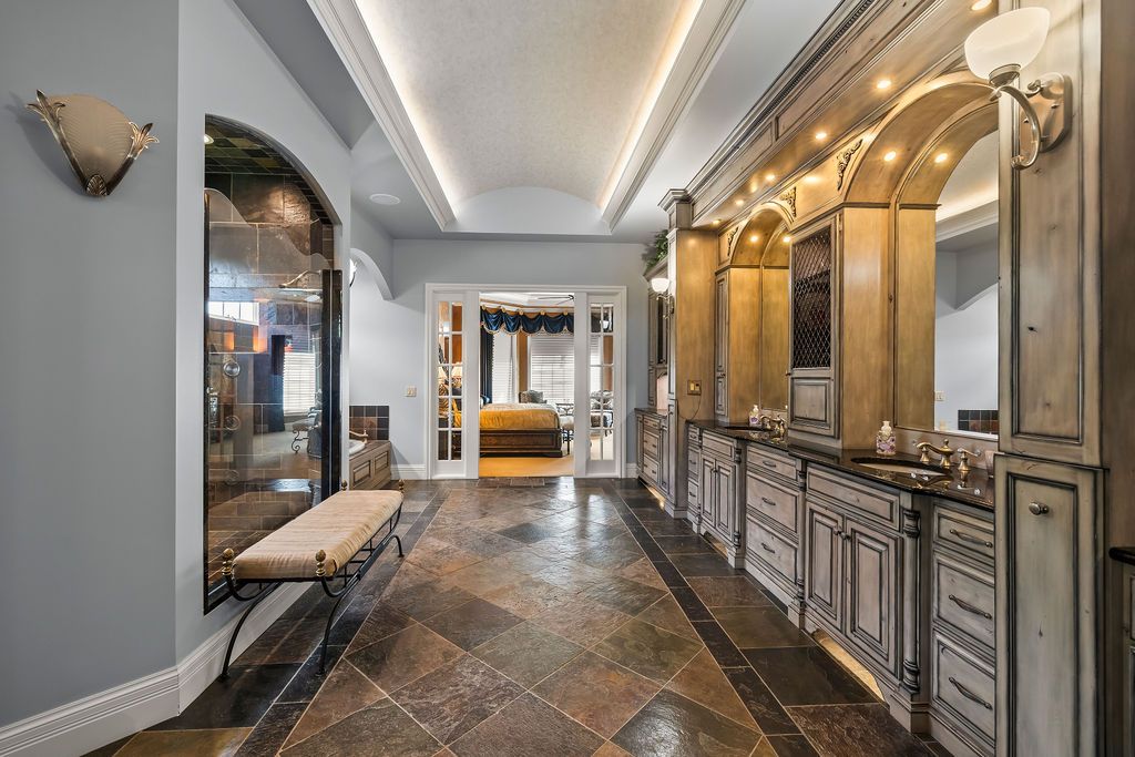 Elegant bathroom hallway with ornate wood cabinetry, arched mirrors, tray ceiling, and dark stone tile flooring.