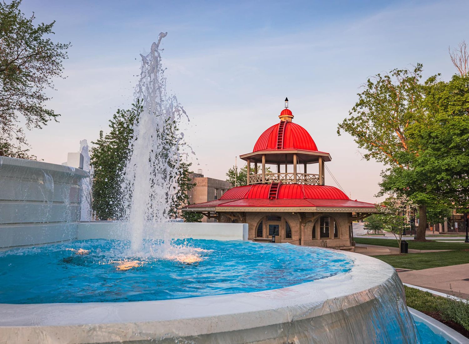 Elegant fountain with water jets in front of a red-domed gazebo pavilion in a scenic city park.