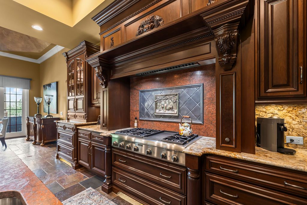 Luxurious kitchen with dark cherry wood cabinetry, granite countertops, professional gas range, and decorative tile backsplash.