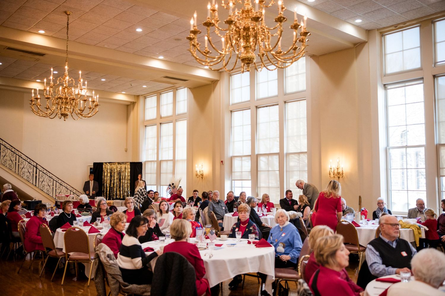 Elegant banquet hall filled with guests in red attire seated at round tables beneath ornate chandeliers.