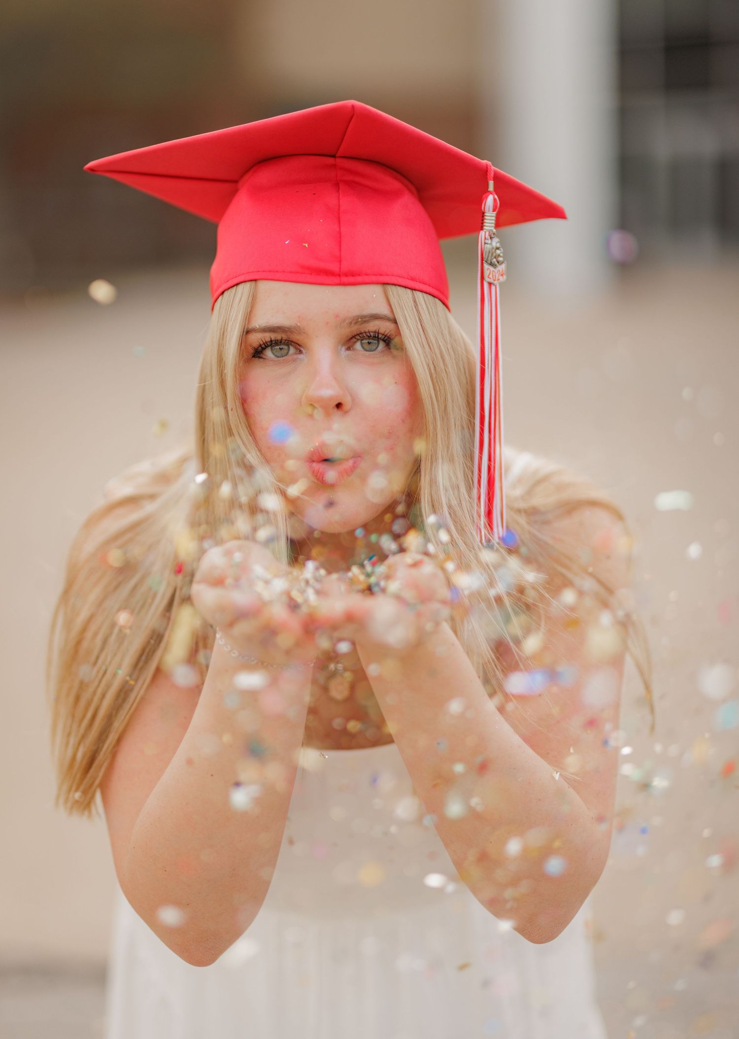 Blonde graduate blowing glitter confetti wearing red cap and white dress in celebratory senior photo.