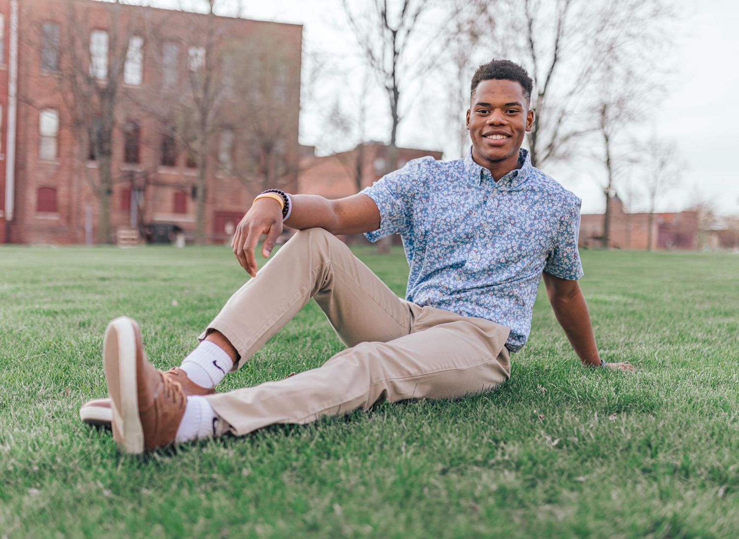 Young man smiling and relaxing on green grass, wearing a floral shirt and khaki pants, with a brick building in the background.