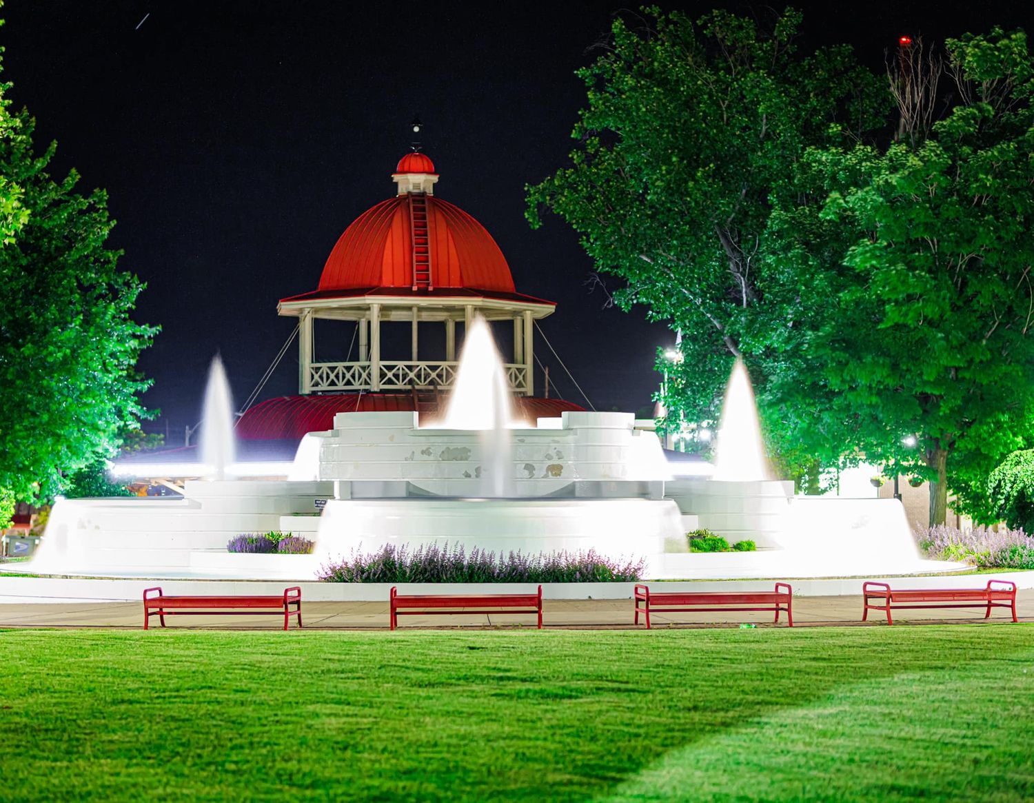 Iconic red-domed gazebo with illuminated fountains and lush green trees glowing at night in a park.