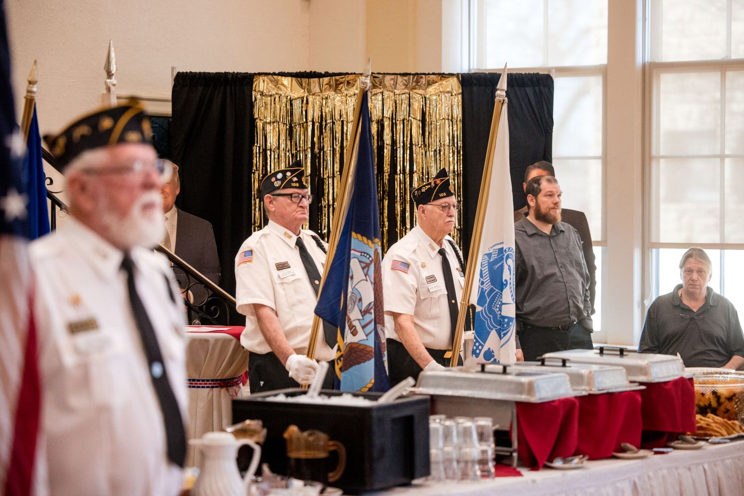 Veterans in uniform holding flags at a formal military ceremony with a decorated backdrop and banquet table.