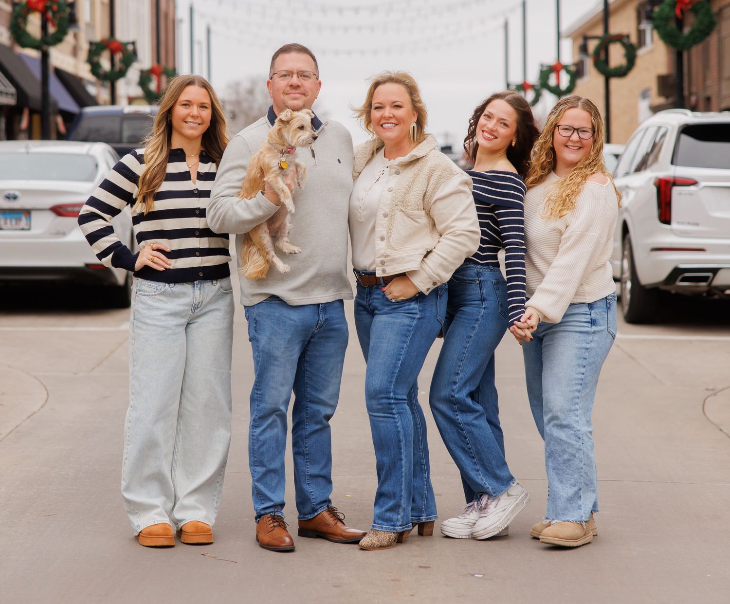 Family of five with dog posing outdoors near festive holiday wreaths on a decorated street.