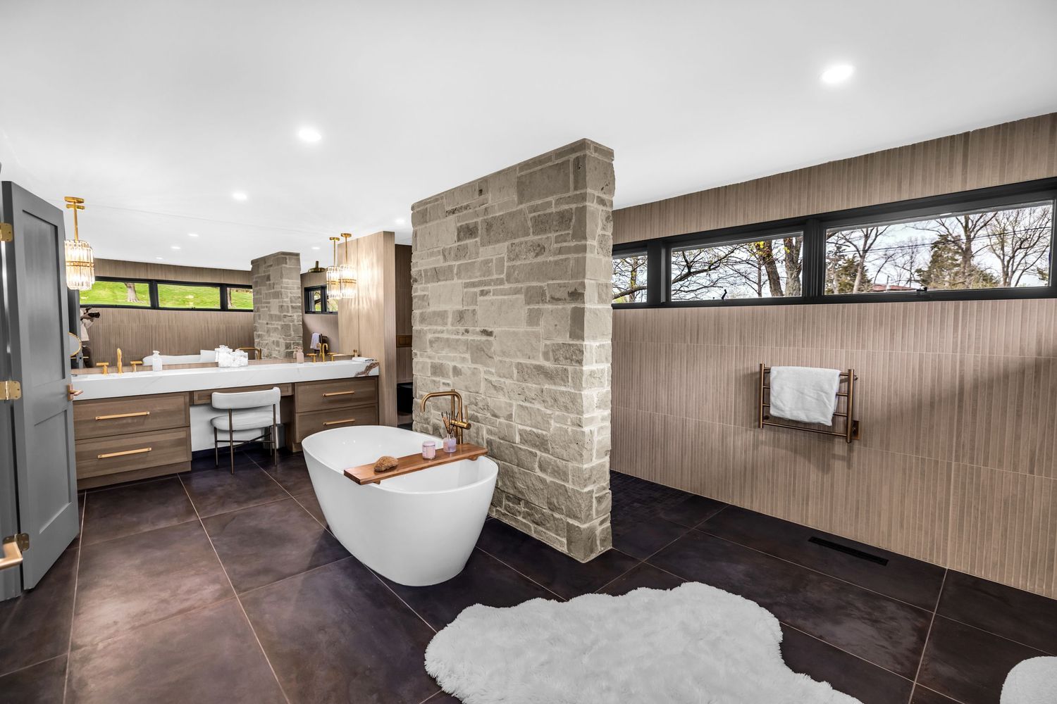 Luxury bathroom featuring freestanding soaking tub, stone brick accent wall, and dark tile flooring.