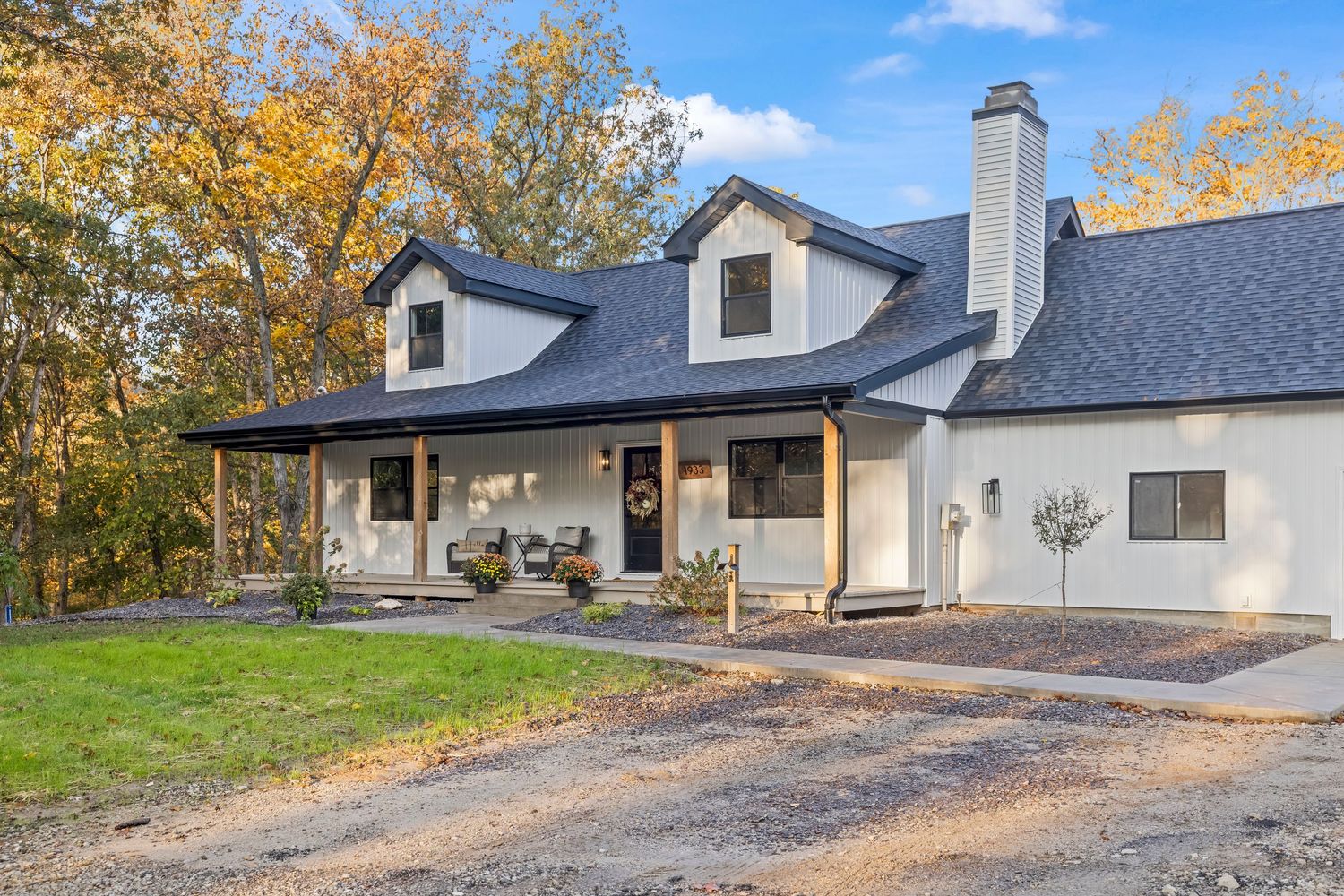 White farmhouse with dark roof, covered porch, and chimney surrounded by autumn trees on a gravel driveway.
