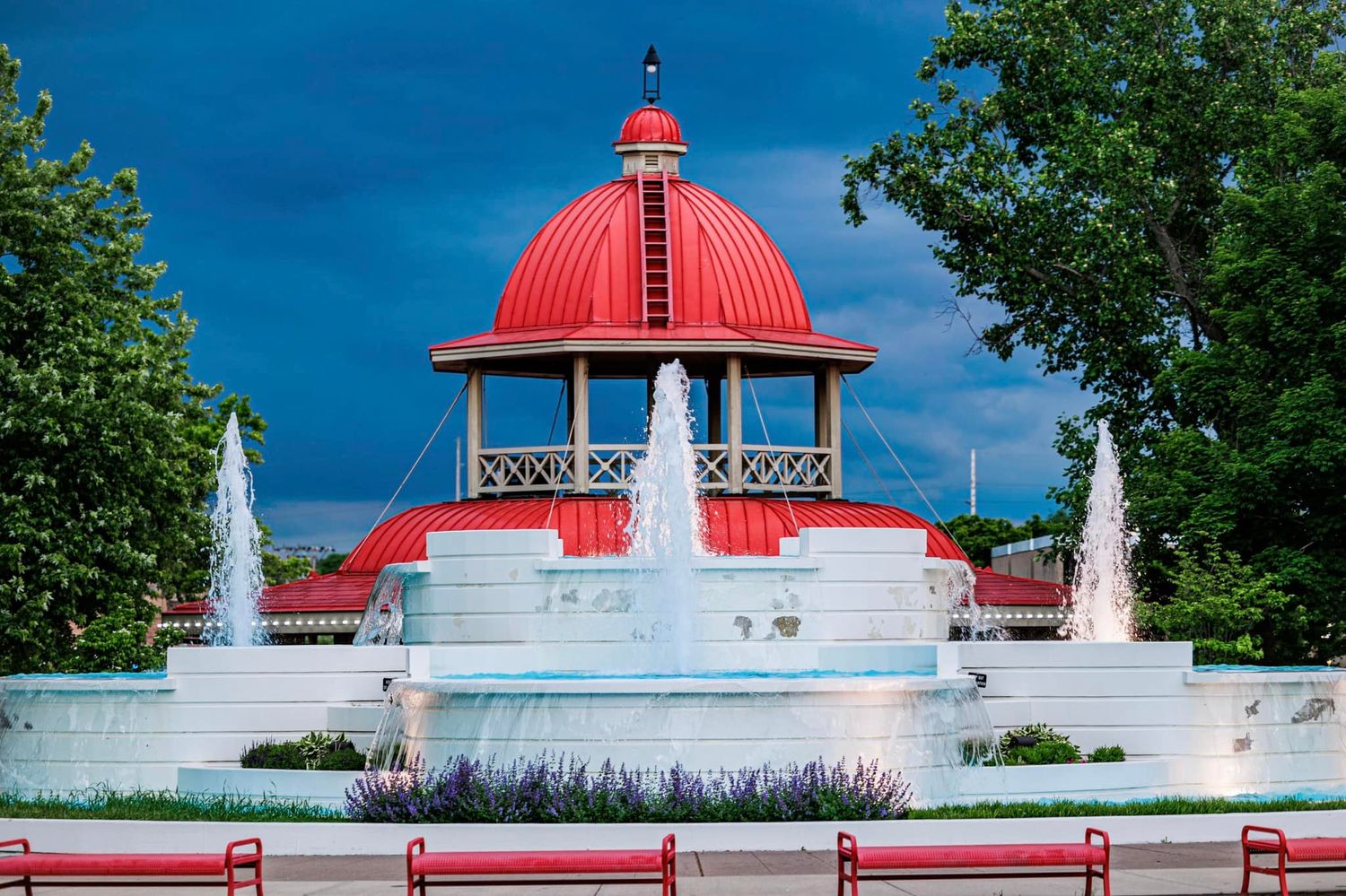 Stunning red-domed gazebo surrounded by symmetrical fountains and red benches under a dramatic cloudy sky.