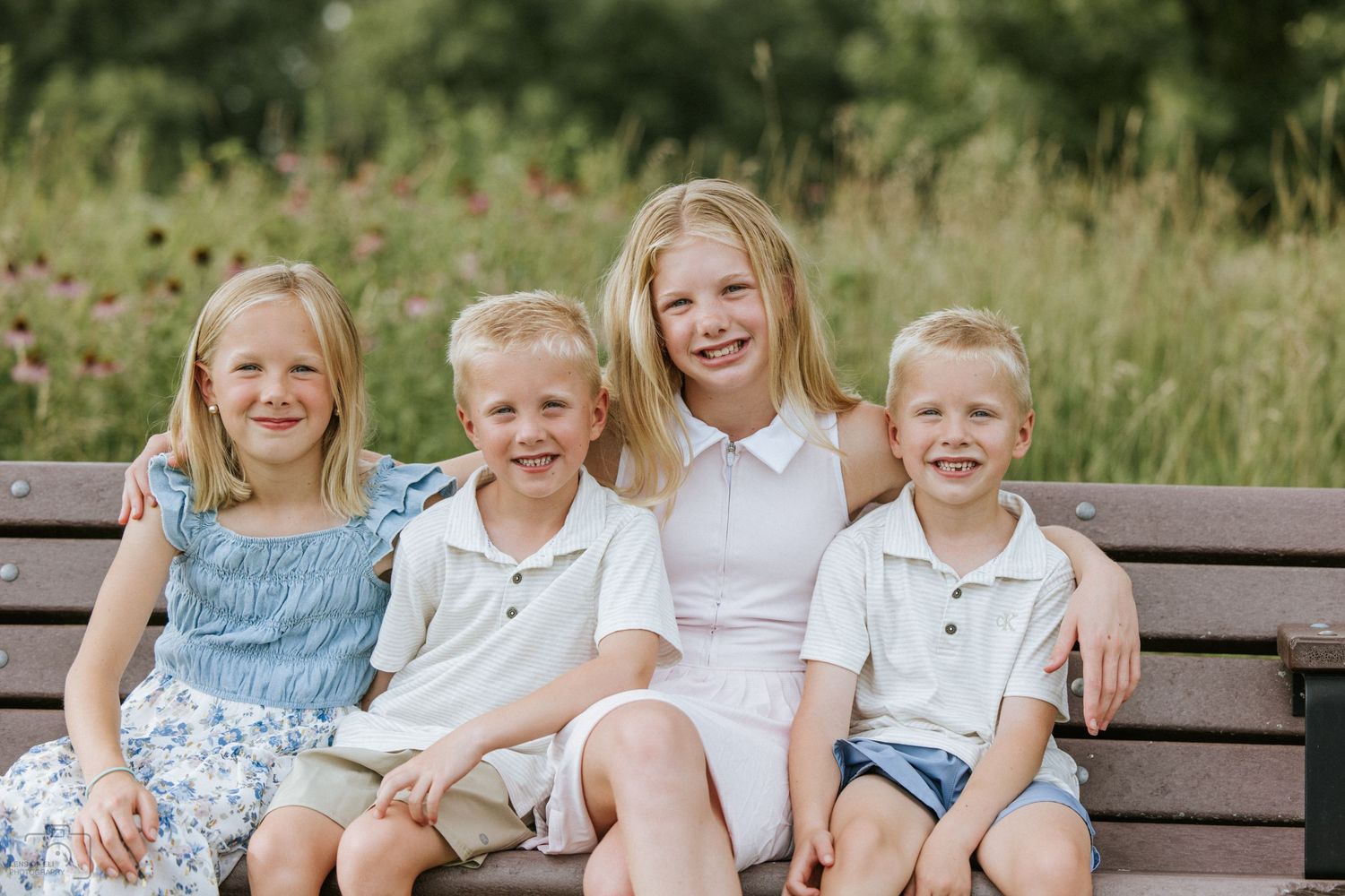 Four blonde siblings smiling together on a wooden bench in a green outdoor setting.
