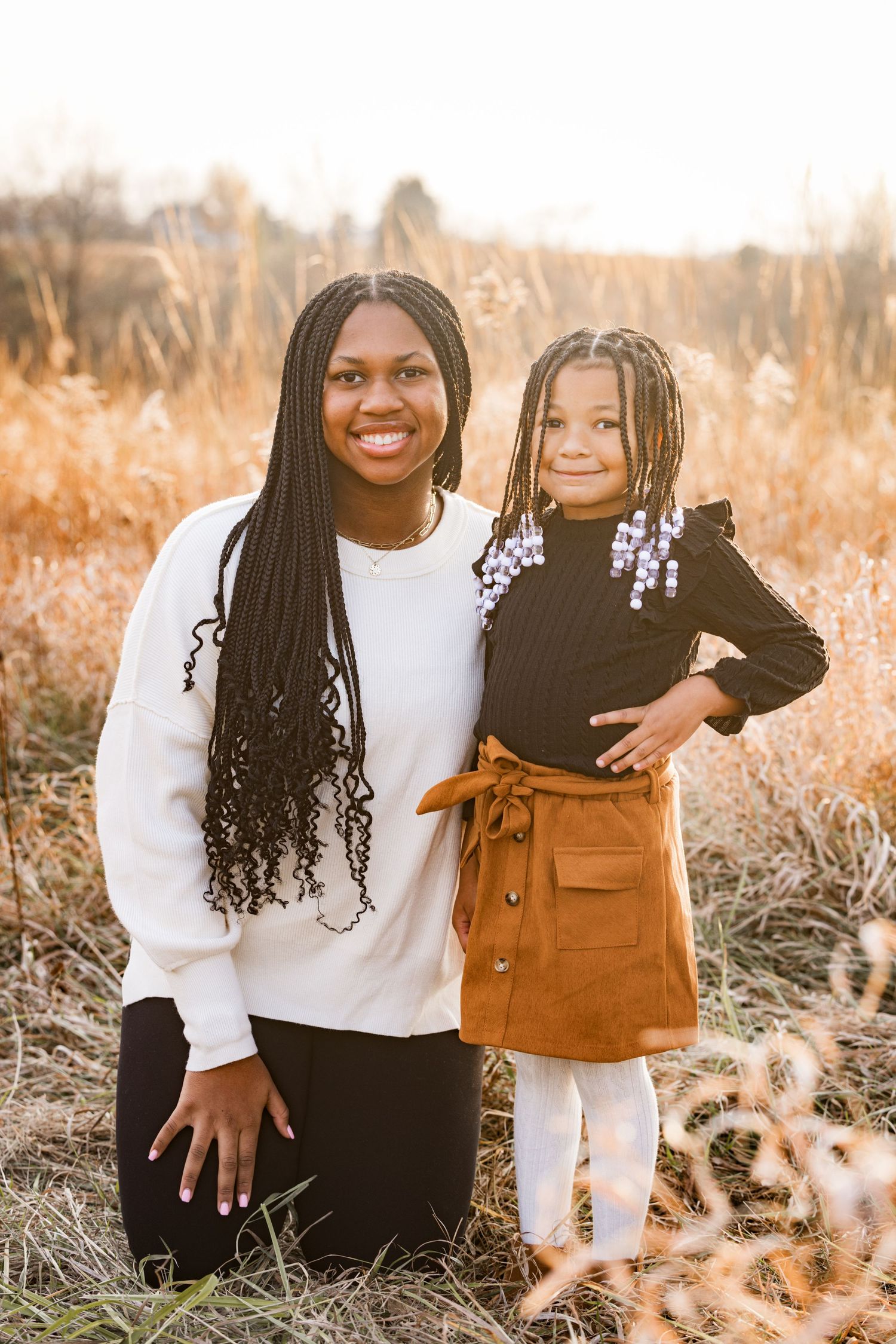 Smiling woman kneeling beside young girl in autumn field during golden hour family portrait.