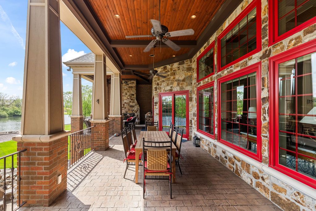 Covered patio with ceiling fan, stone columns, outdoor dining set, and red-framed windows on a luxury home.