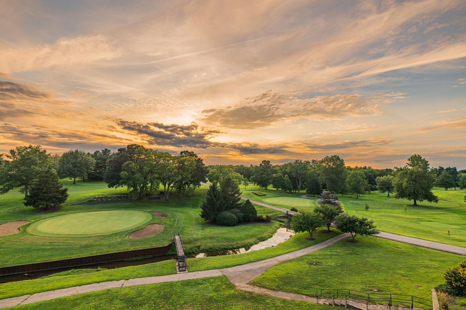 Aerial sunset view of lush green golf course with winding paths and trees under a colorful sky.