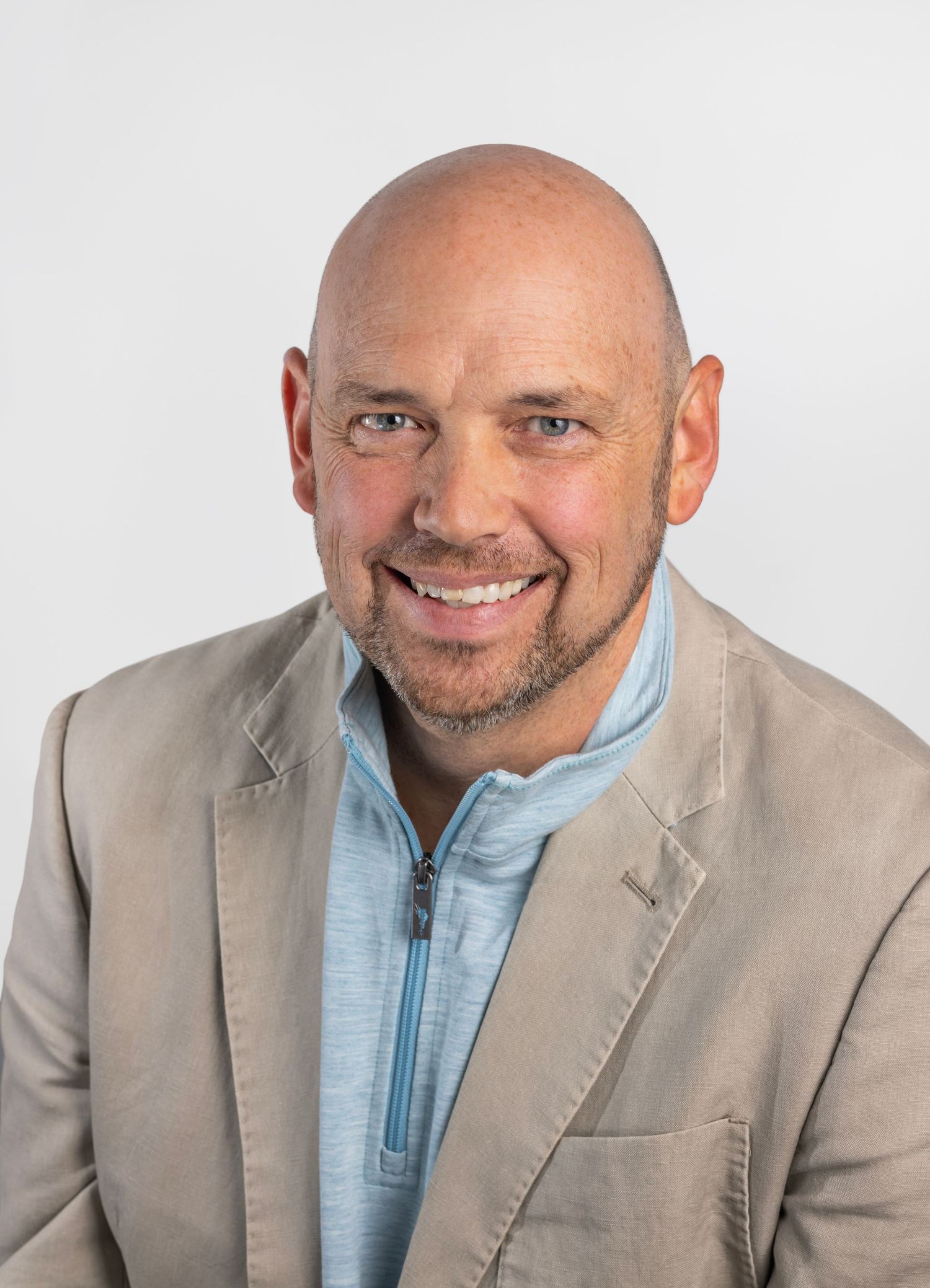 Smiling man in beige blazer and blue zip shirt poses for professional headshot.