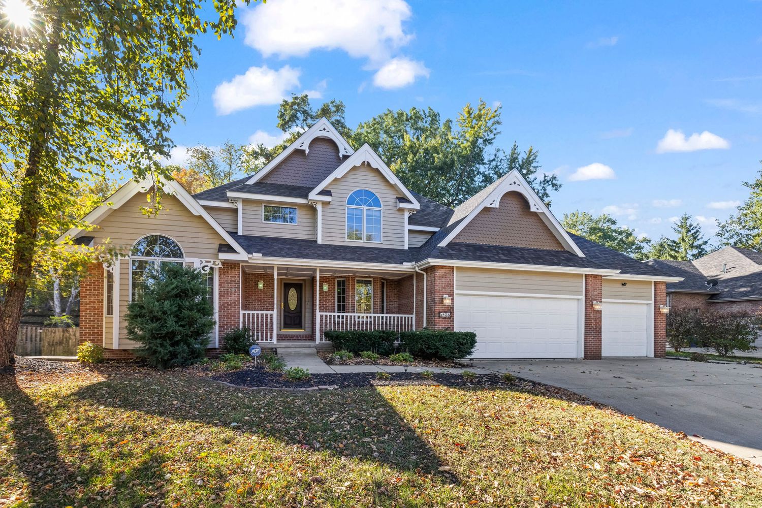 Two-story brick home with attached two-car garage, arched window, covered porch, and mature trees on a sunny autumn day.