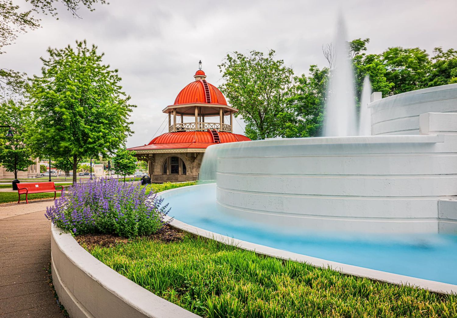 White fountain with blue water in a park beside a red-domed gazebo surrounded by purple flowers.