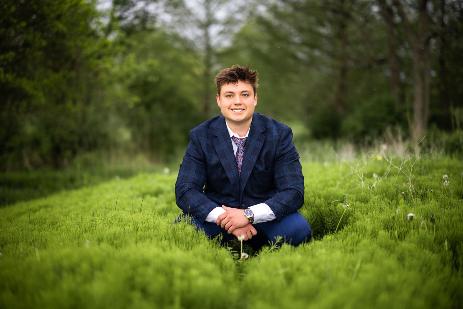 Young man in navy suit sitting relaxed in a lush green grassy outdoor setting.