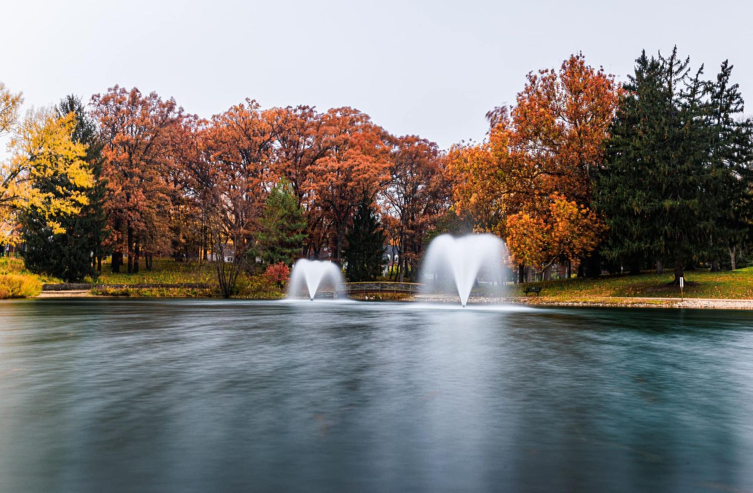 Two elegant water fountains spraying in a calm pond surrounded by vibrant autumn foliage in a park.