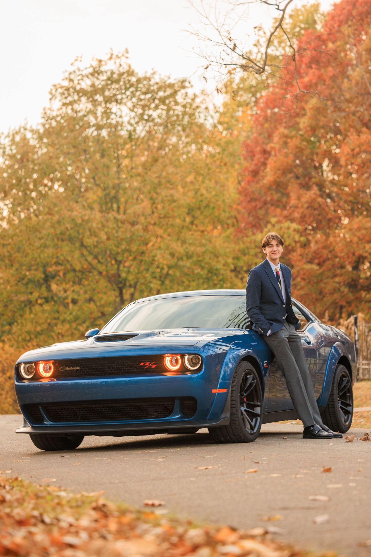 Teen boy in suit leaning on blue Dodge Challenger among fall foliage for senior portrait.