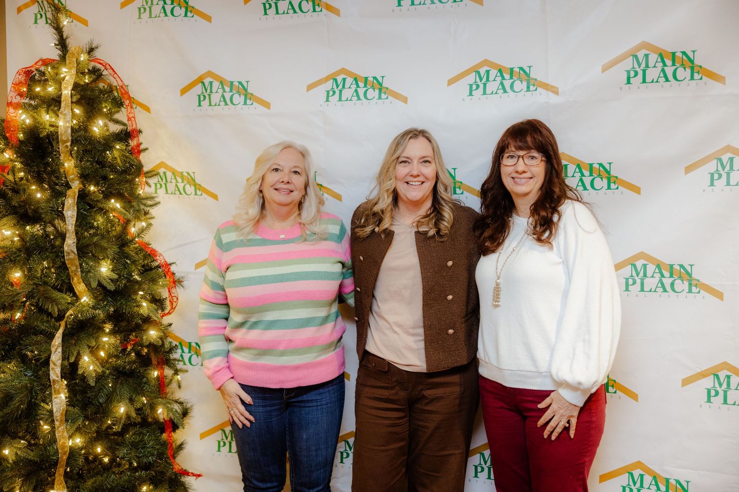 Three women smiling together in front of a Main Place branded backdrop near a decorated Christmas tree.
