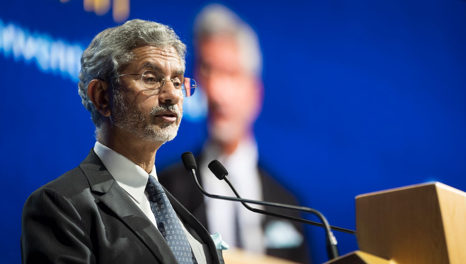 A distinguished man in a suit speaks at a podium with microphones at a formal conference event.