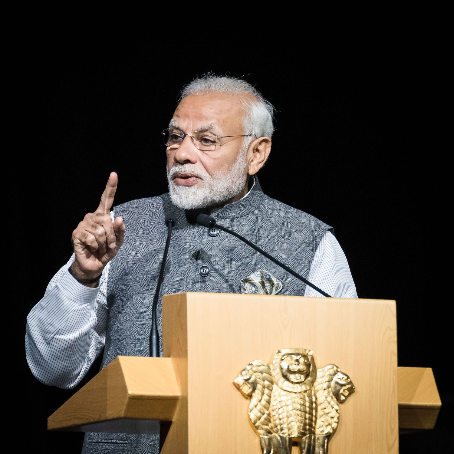 A distinguished elderly man in a gray vest speaks at a podium with the Indian national emblem during an official address.