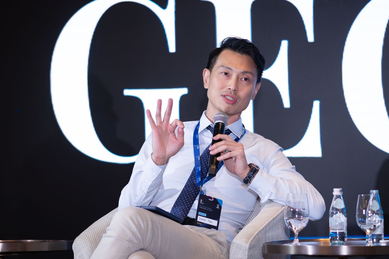 Young man in white shirt and tie gesturing during conference panel discussion.