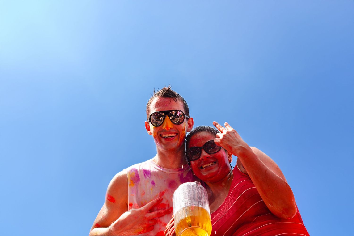 Two smiling friends covered in colorful powder pose together under a bright blue sky at a Holi festival.