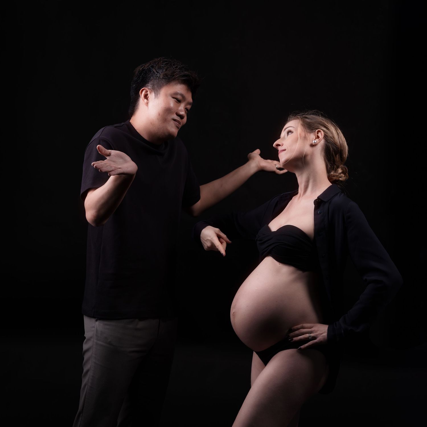 A pregnant woman in black maternity wear poses with a man against a dark studio background.