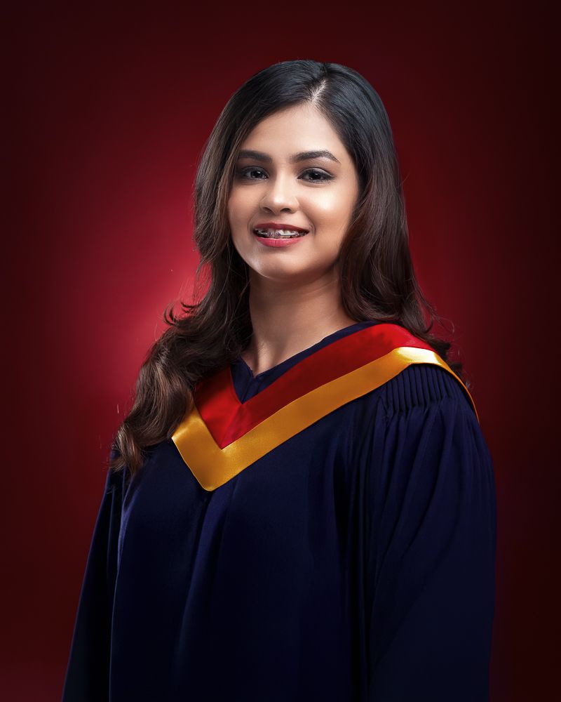 A young woman in a navy blue graduation gown with red and gold hood smiles against a red background.