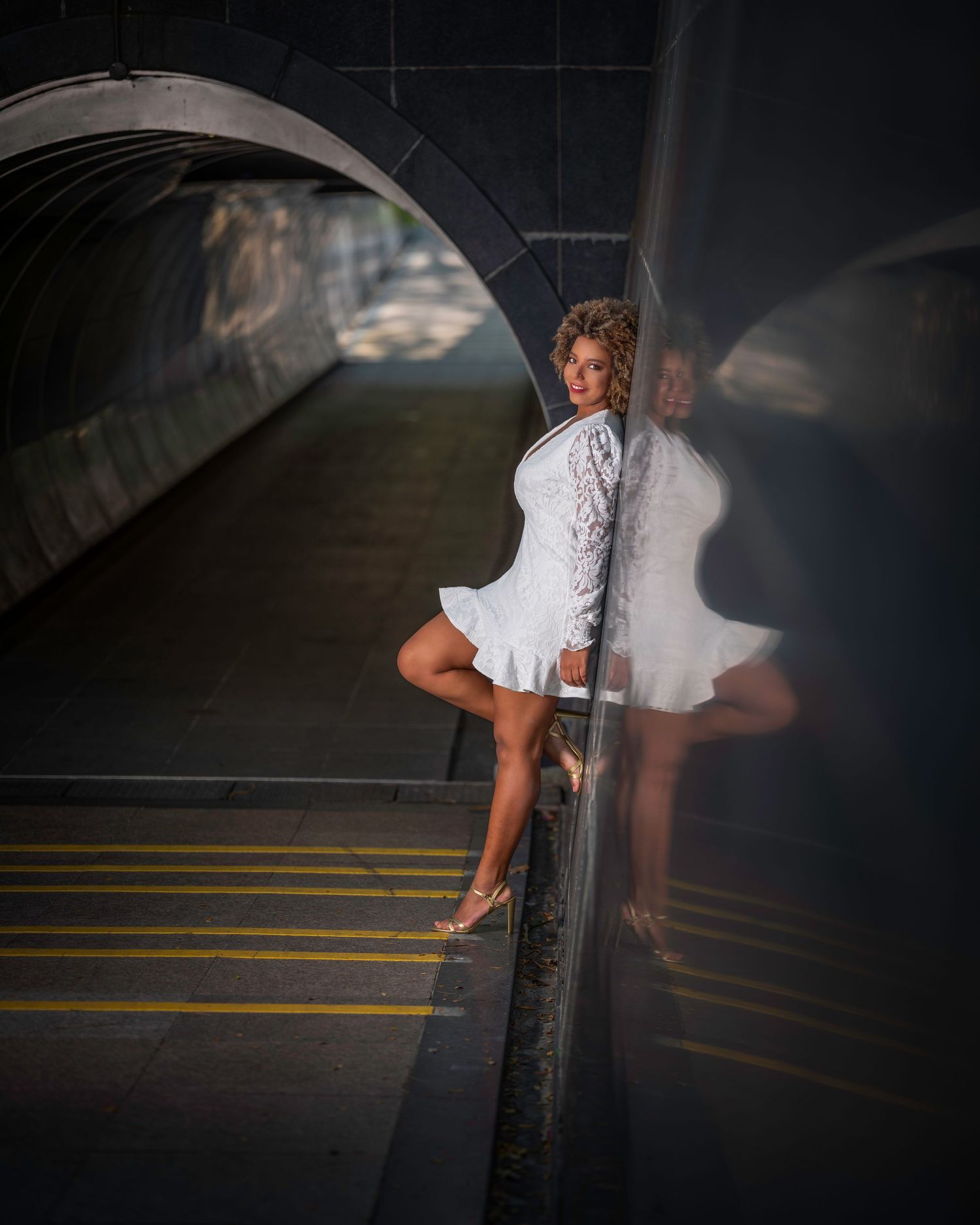 Woman in white mini dress posing against reflective dark pillar in urban tunnel setting.