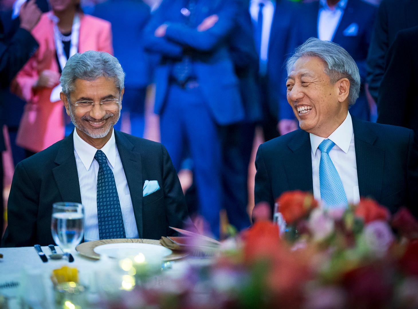 Two men in dark suits laughing together at a formal dinner event with floral arrangements.