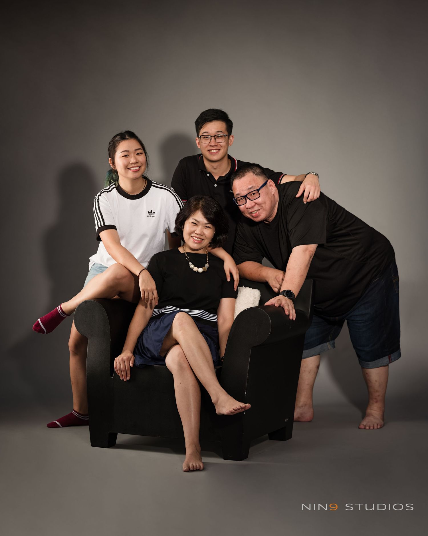 Family of four posing together in a studio portrait, three seated and one standing, smiling warmly at the camera.