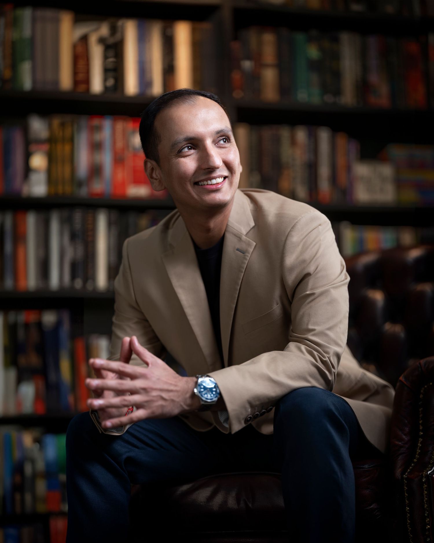A smiling man in a beige blazer sits confidently in front of a bookshelf in a professional portrait photo.