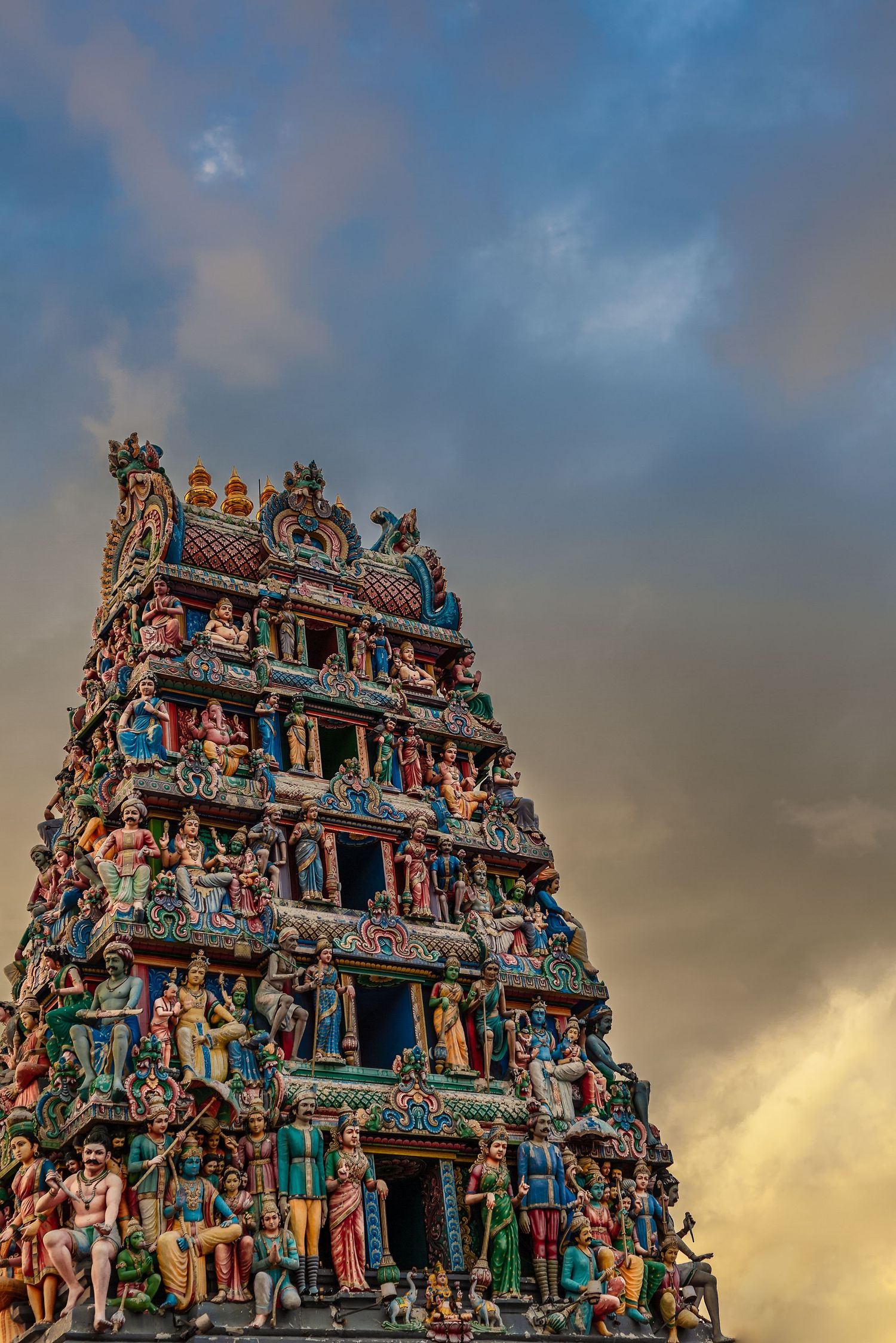 Colorful Hindu temple gopuram adorned with intricate sculptures of deities against a dramatic cloudy sky.