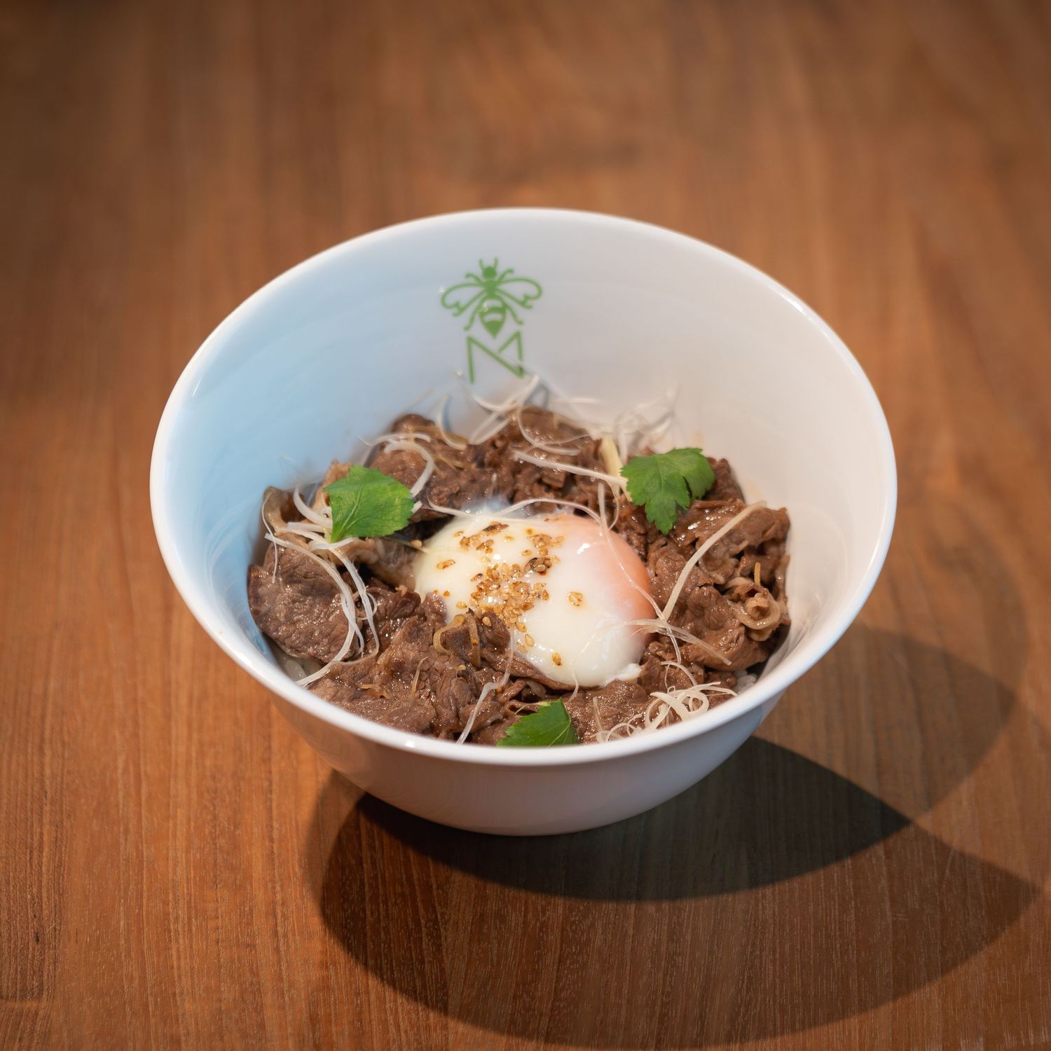 A white ceramic bowl filled with braised beef, a soft-boiled egg, sprouts, and fresh herbs on a wooden table.