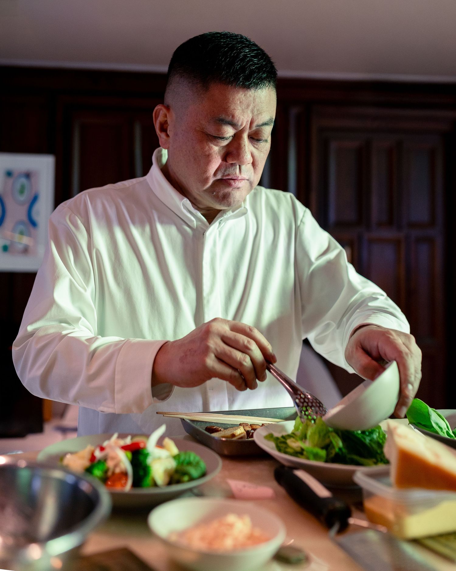 A chef in a white shirt carefully plates a gourmet dish surrounded by fresh vegetables and ingredients on a wooden table.