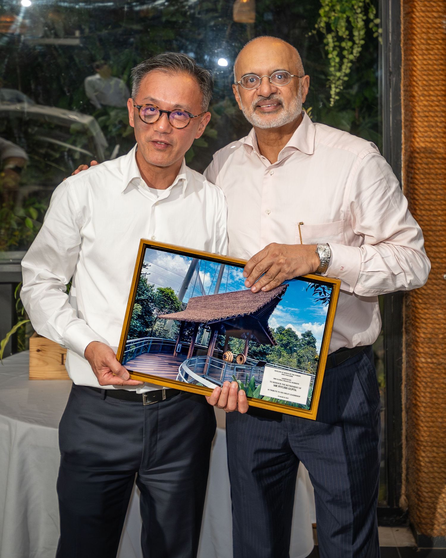 Two men in white shirts hold a framed photo of a traditional pavilion at a formal recognition ceremony.