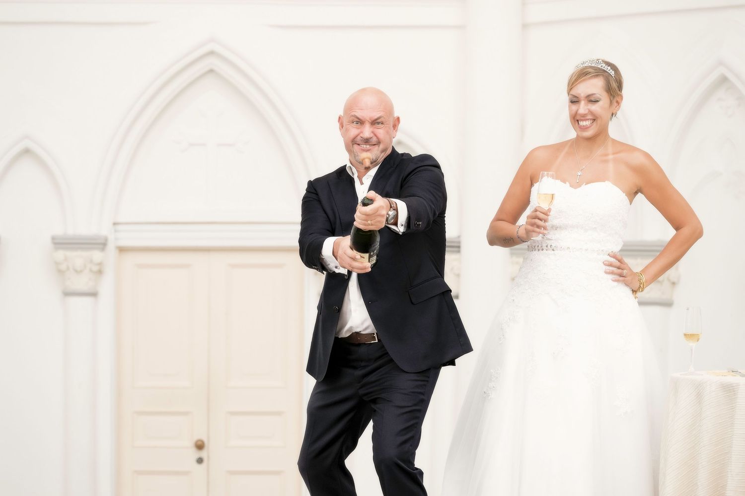 Bald groom popping champagne bottle beside laughing bride in strapless gown in white chapel.