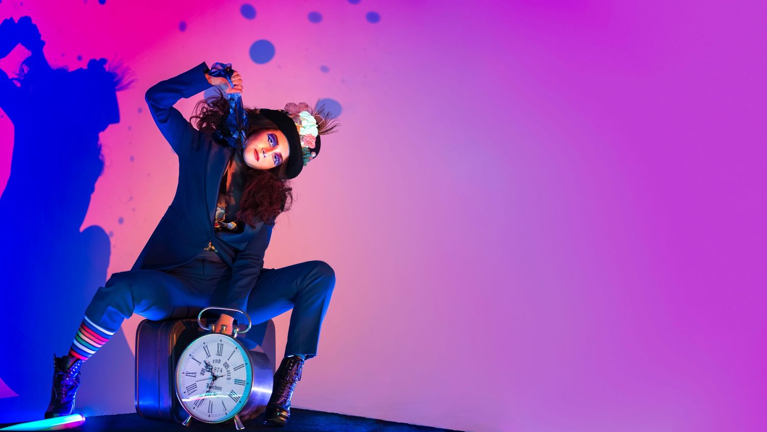 A woman in blue denim poses dynamically beside a large clock under vibrant pink and blue neon lights.