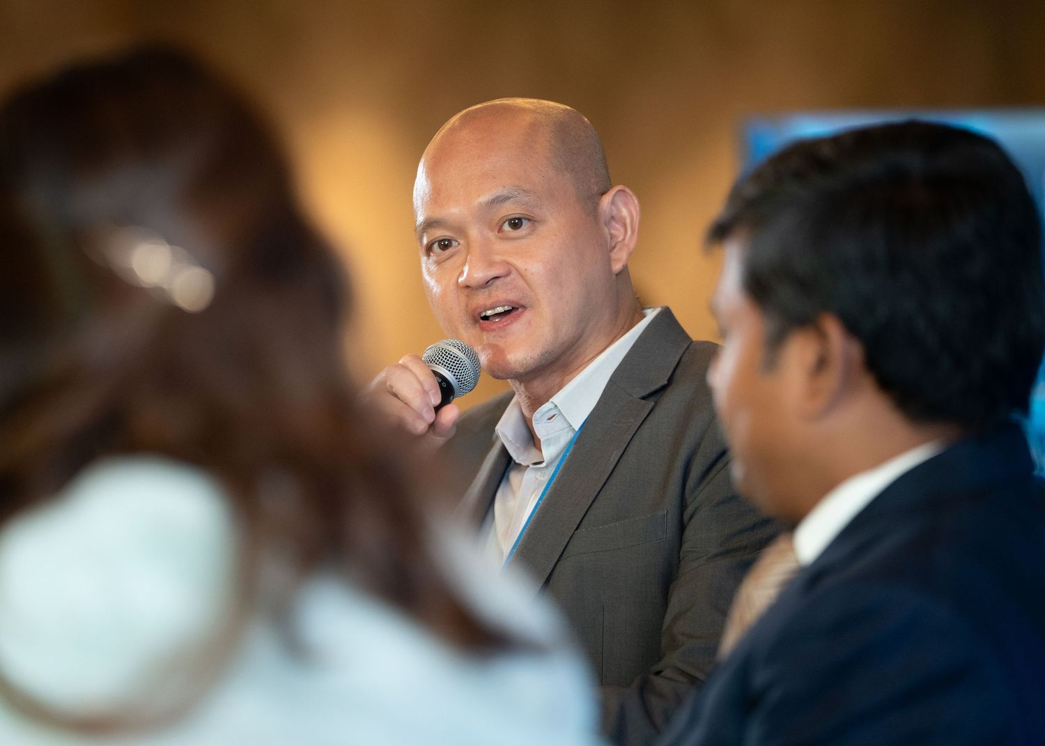 A man in a gray blazer speaks into a microphone during a panel discussion at a professional conference.