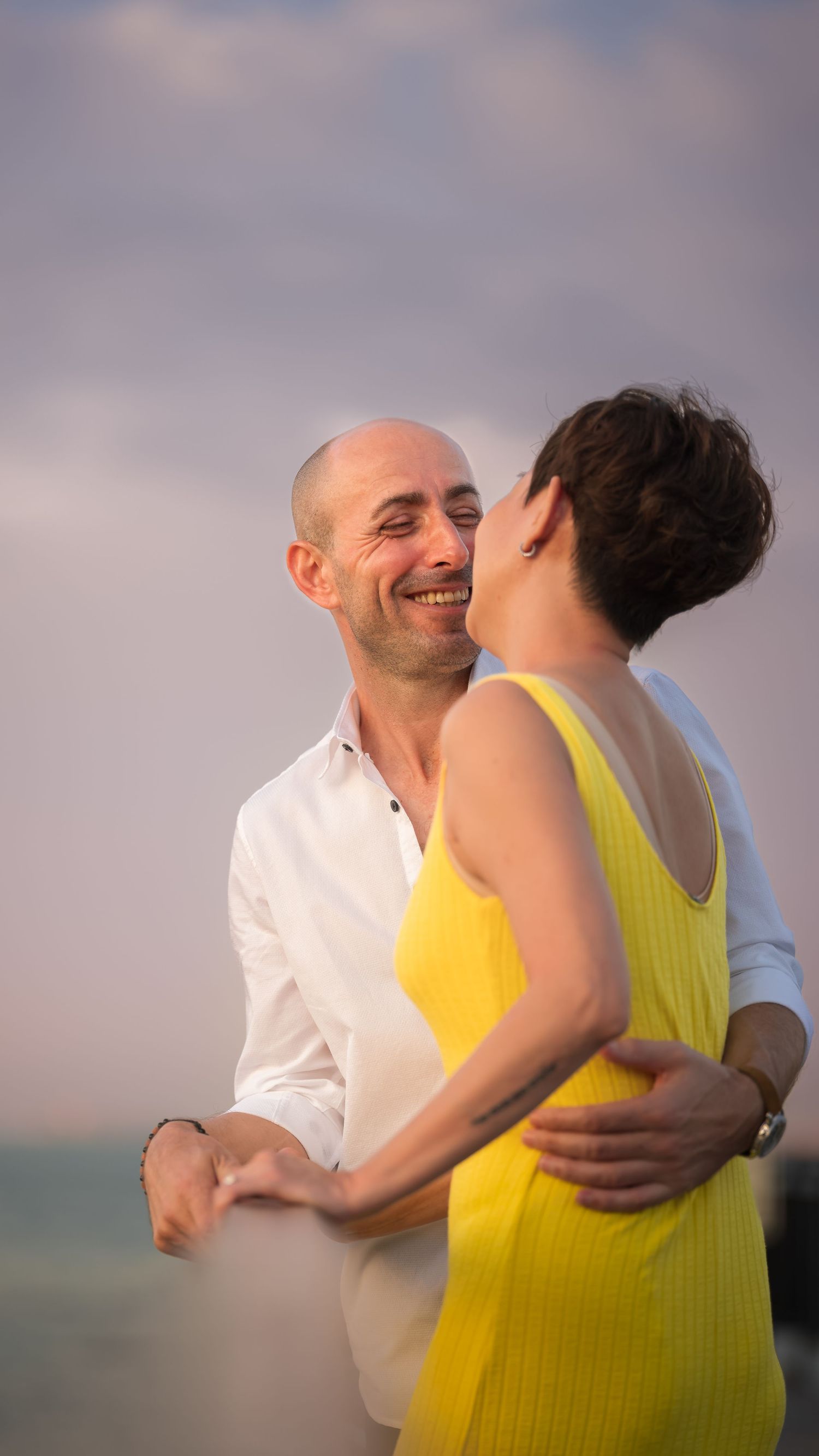 Happy bald man in white shirt embracing woman in yellow dress, sharing a joyful moment at sunset by the ocean.
