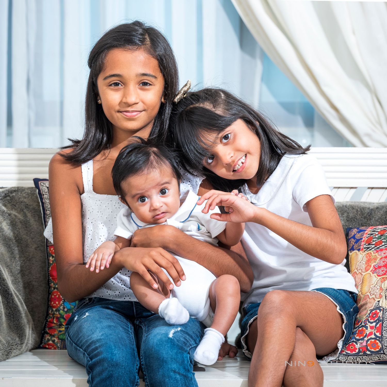 Three young siblings pose together on a sofa, two older girls smiling with a baby in white outfit.